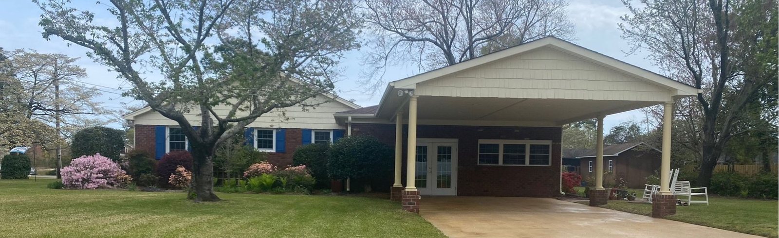 Brick ranch with a carport and a clean concrete driveway, azaleas, and spring trees.